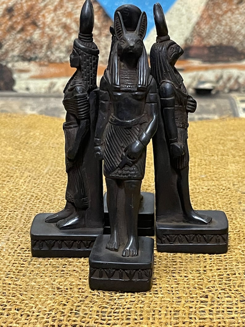 May include: Three black figurines of Egyptian gods, each standing on a square base with intricate carvings. The god in the center is Anubis, the jackal-headed god of the dead, with a headdress and a staff. The god on the left is a woman with a tall headdress and a staff. The god on the right is a man with a falcon head and a staff.