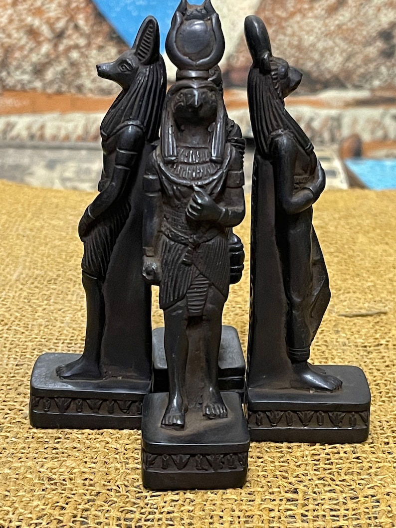 May include: Three black statues of Egyptian deities. The statues are standing on small square bases. The statue on the left is a jackal-headed god, the middle statue is a falcon-headed god, and the statue on the right is a rabbit-headed god.