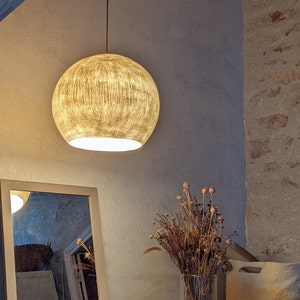 Puede incluir: Una l&aacute;mpara colgante redonda y texturizada con un brillo dorado y c&aacute;lido cuelga de un cable marr&oacute;n. La habitaci&oacute;n presenta una paleta de colores neutros, que incluye un espejo, una cesta tejida y un jarr&oacute;n con flores secas.