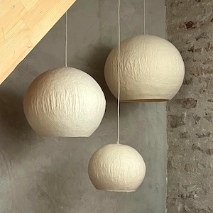 May include: Three off-white, spherical pendant lights of varying sizes hang from the ceiling. The textured surface of each light fixture contrasts with the smooth, neutral-toned wall and wooden beam in the background. A woven basket sits below.