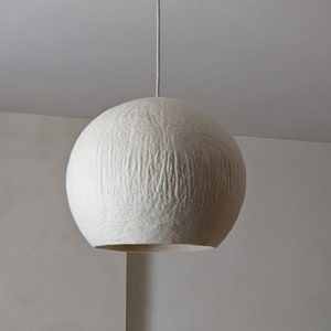 Puede incluir: Una l&aacute;mpara colgante esf&eacute;rica blanca. La pantalla texturizada cuelga de un soporte de techo blanco mediante un cable beige. La l&aacute;mpara tiene forma redonda y una superficie texturizada, creando un brillo suave y difuso.