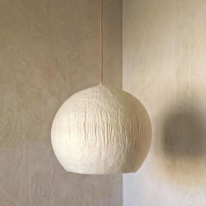 Puede incluir: Una l&aacute;mpara colgante esf&eacute;rica de color blanquecino con una superficie texturizada, suspendida de un cable marr&oacute;n. La luz cuelga en la esquina de una habitaci&oacute;n con paredes de tonos neutros.