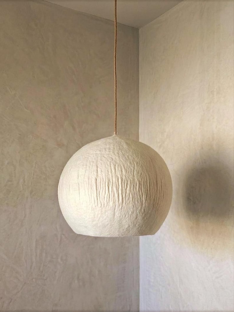 Puede incluir: Una l&aacute;mpara colgante esf&eacute;rica de color blanquecino con una superficie texturizada, suspendida de un cable marr&oacute;n. La luz cuelga en la esquina de una habitaci&oacute;n con paredes de tonos neutros.
