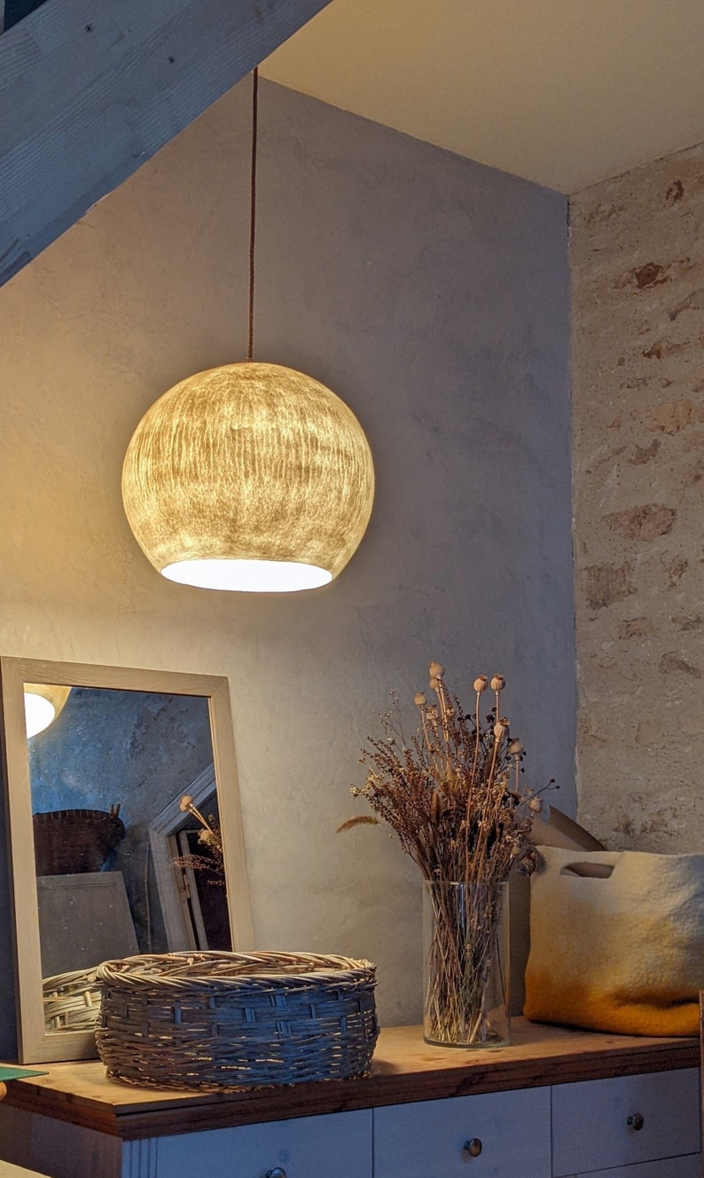 Puede incluir: Una l&aacute;mpara colgante redonda y texturizada con un brillo dorado y c&aacute;lido cuelga de un cable marr&oacute;n. La habitaci&oacute;n presenta una paleta de colores neutros, que incluye un espejo, una cesta tejida y un jarr&oacute;n con flores secas.