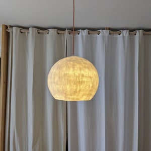 Puede incluir: Una l&aacute;mpara colgante esf&eacute;rica con una pantalla texturizada de color blanquecino. La luz cuelga de un cable marr&oacute;n y un soporte de techo blanco. El fondo presenta cortinas blancas y un marco de madera.