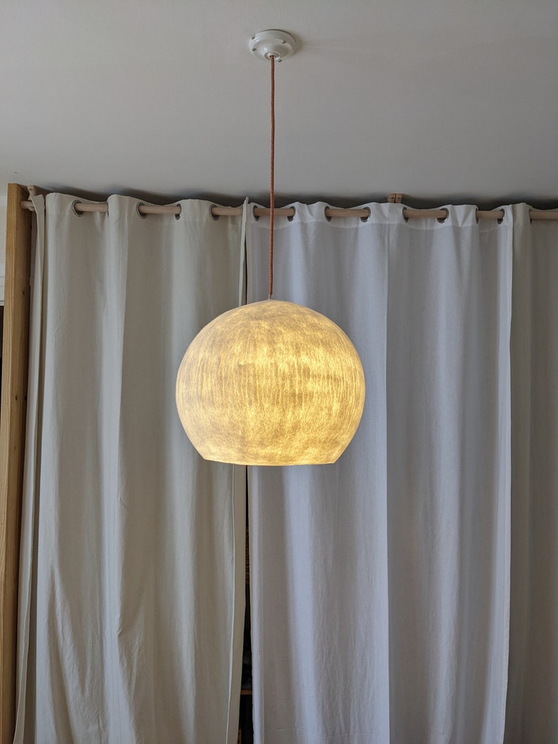 Puede incluir: Una l&aacute;mpara colgante esf&eacute;rica con una pantalla texturizada de color blanquecino. La luz cuelga de un cable marr&oacute;n y un soporte de techo blanco. El fondo presenta cortinas blancas y un marco de madera.