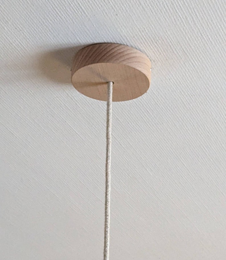 Puede incluir: Un dosel de techo de madera clara, de dise&ntilde;o redondo. Un cable beige se extiende desde el centro del dosel, colgando hacia abajo. El dosel est&aacute; montado en un techo blanco.