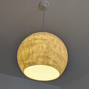 Puede incluir: Una l&aacute;mpara colgante esf&eacute;rica con una pantalla beige texturizada. La luz cuelga de un soporte de techo blanco y un cable delgado. La parte inferior de la pantalla est&aacute; abierta, revelando una luz c&aacute;lida y brillante.