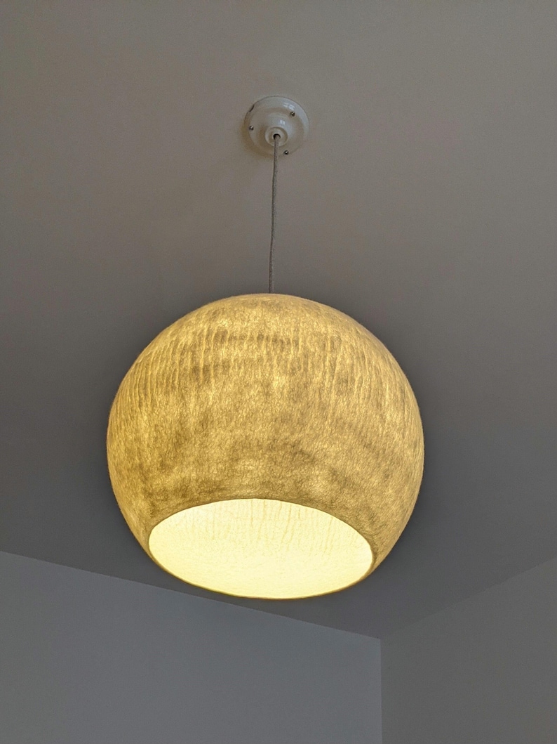Puede incluir: Una l&aacute;mpara colgante esf&eacute;rica con una pantalla beige texturizada. La luz cuelga de un soporte de techo blanco y un cable delgado. La parte inferior de la pantalla est&aacute; abierta, revelando una luz c&aacute;lida y brillante.