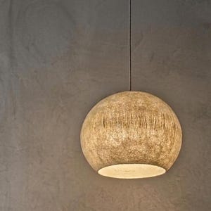 Puede incluir: Una l&aacute;mpara colgante esf&eacute;rica con una superficie beige texturizada. La luz cuelga de un fino cable negro sobre un fondo gris neutro. La parte inferior de la luz emite un brillo c&aacute;lido.