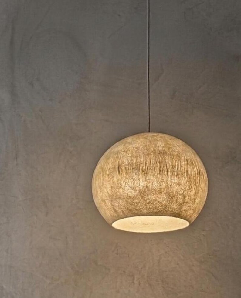 Puede incluir: Una l&aacute;mpara colgante esf&eacute;rica con una superficie beige texturizada. La luz cuelga de un fino cable negro sobre un fondo gris neutro. La parte inferior de la luz emite un brillo c&aacute;lido.