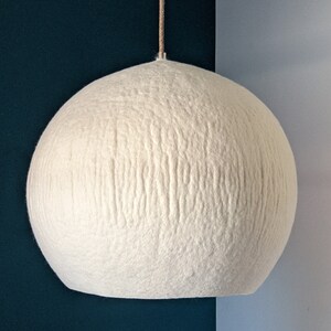 Puede incluir: Una l&aacute;mpara colgante esf&eacute;rica grande, de color blanquecino. La superficie texturizada de la l&aacute;mpara est&aacute; hecha de un material suave, similar al fieltro. La luz cuelga de un fino cord&oacute;n beige sobre un fondo azul oscuro y azul claro.