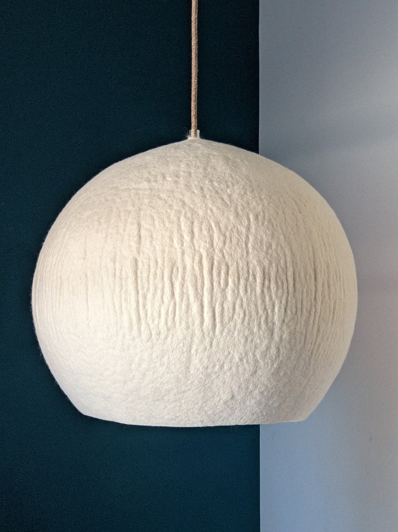 Puede incluir: Una l&aacute;mpara colgante esf&eacute;rica grande, de color blanquecino. La superficie texturizada de la l&aacute;mpara est&aacute; hecha de un material suave, similar al fieltro. La luz cuelga de un fino cord&oacute;n beige sobre un fondo azul oscuro y azul claro.
