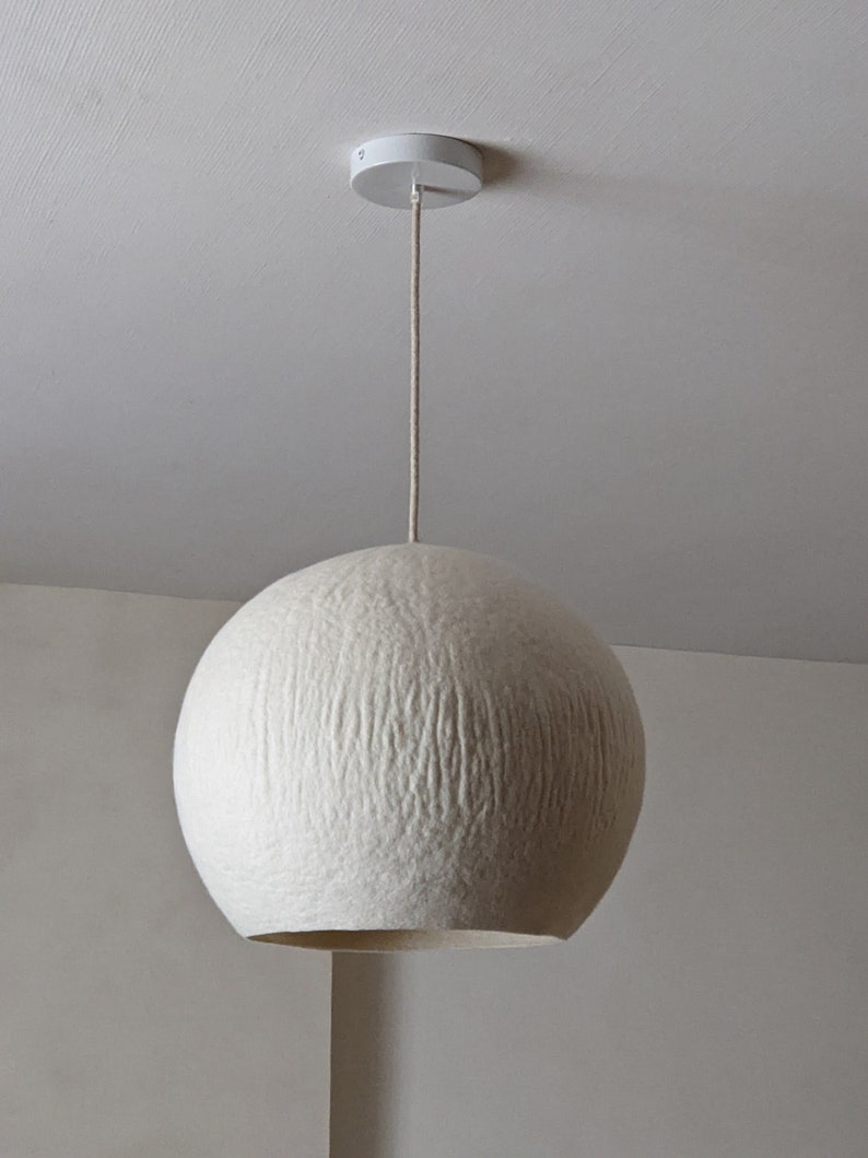 Puede incluir: Una l&aacute;mpara colgante esf&eacute;rica blanca. La pantalla texturizada cuelga de un soporte de techo blanco mediante un cable beige. La l&aacute;mpara tiene forma redonda y una superficie texturizada, creando un brillo suave y difuso.