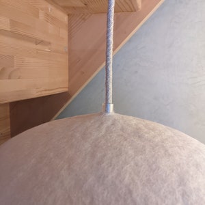 Puede incluir: Una l&aacute;mpara colgante de fieltro rosa claro con un accesorio de metal plateado y un cable trenzado gris. La l&aacute;mpara tiene una forma redondeada, similar a una c&uacute;pula. El fondo incluye vigas de madera y una pared azul claro.