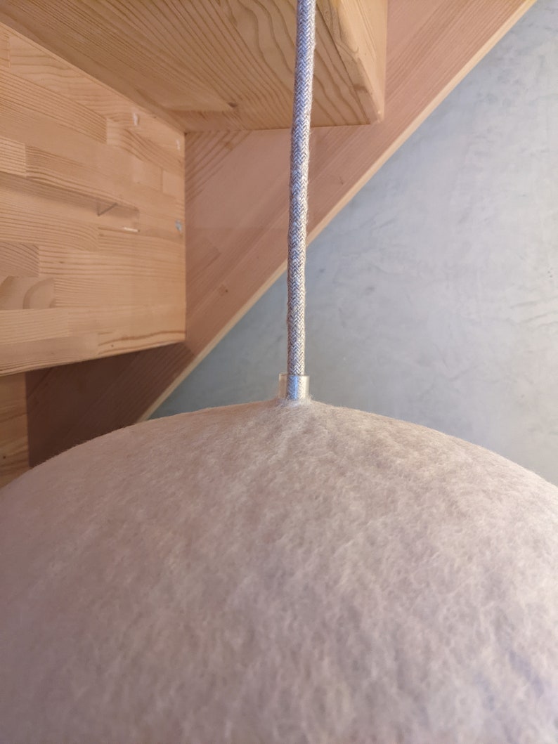 Puede incluir: Una l&aacute;mpara colgante de fieltro rosa claro con un accesorio de metal plateado y un cable trenzado gris. La l&aacute;mpara tiene una forma redondeada, similar a una c&uacute;pula. El fondo incluye vigas de madera y una pared azul claro.
