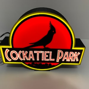 May include: A black and yellow sign with a red background and a silhouette of a cockatiel bird. The sign reads "Cockatiel Park".