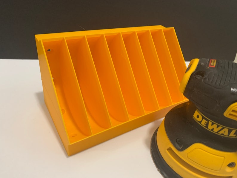 K&ouml;nnte beinhalten: Ein gelber Plastik-Organizer mit sechs F&auml;chern zum Verstauen von Schleifscheiben. Im Vordergrund befindet sich eine schwarze und gelbe DeWalt-Schleifmaschine.