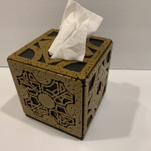 May include: A gold-colored tissue box with intricate designs, resembling a puzzle box. The box has a black interior and a white tissue dispenser.