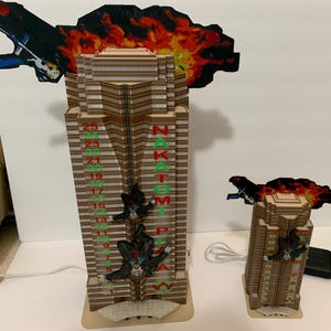 May include: Two model buildings, one larger than the other, depicting the Nakatomi Plaza from the movie Die Hard. The larger building has a red and green sign that reads "Nakatomi Plaza" and has a fire explosion above it. The smaller building has a similar fire explosion above it. Both buildings have figures hanging from ropes.