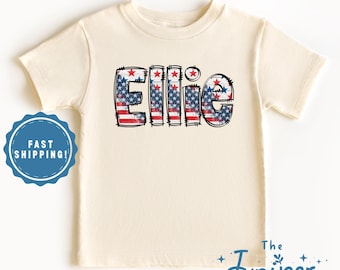May include: Cream-colored toddler t-shirt with the name "Ellie" in a patriotic design. The letters are filled with a red, white, and blue American flag pattern, outlined in black. The shirt is short-sleeved and has a round neckline.