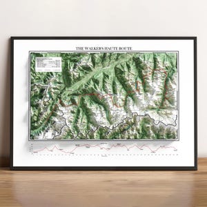 May include: Framed print of The Walkers Haute Route, a detailed map of a mountain range with contour lines, elevation data, and a red trail line. The print is in a black frame and rests on a wooden surface.