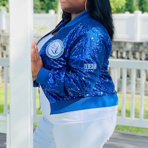 May include: A royal blue sequined bomber jacket with a white top and white pants. The jacket has a circular patch with text and the year "1920" on the sleeve. The jacket has blue ribbed cuffs and a matching collar.
