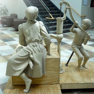 May include: Two clay sculptures of a girl and a boy, the girl is sitting on a wooden platform and the boy is standing next to a hand pump, both sculptures are in a light brown color.