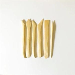 May include: Set of seven wooden sculpting tools with various shapes and sizes. The tools are made of natural wood and have a light brown color.