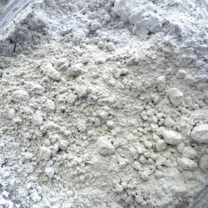May include: Close-up of a container filled with a light gray, powdery substance. The powder has a fine texture with small, irregular clumps throughout. The container's rim is visible, suggesting a wide opening.
