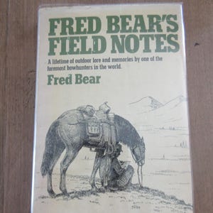 May include: A vintage book titled "Fred Bear's Field Notes" with a drawing of a horse and a person. The cover is tan with green text. The book is about outdoor lore and memories by a bowhunter.