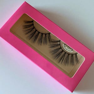 May include: A pair of false eyelashes in a pink box with a clear window. The eyelashes are long and thick, and they are designed to give a dramatic look.