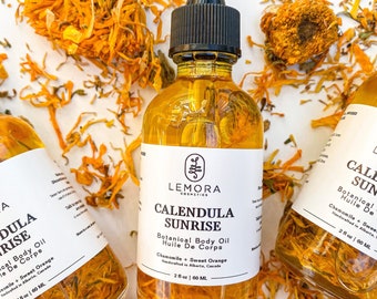 May include: Two amber glass bottles of Calendula Sunrise Botanical Body Oil, with a black dropper top, on a white surface with dried orange flower petals. The label reads "Calendula Sunrise Botanical Body Oil Huile De Corps Chamomile + Sweet Orange Handcrafted in Alberta, Canada 2 fl oz | 60 ML"