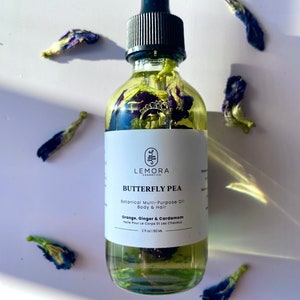 Könnte beinhalten: Eine klare Glasflasche mit schwarzem Tropfer, gefüllt mit einer gelben Flüssigkeit und blauen Schmetterlingserbsenblüten. Das Flaschenetikett lautet "LEMORA COSMETICS BUTTERFLY PEA Botanical Multi-Purpose Oil Body & Hair Orange, Ginger & Cardamom Huile Pour Le Corps Et Les Cheveux 2 fl oz / 60 ML".