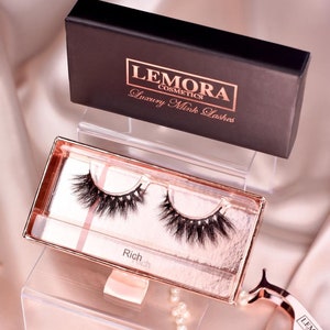 May include: A pair of black mink eyelashes in a clear plastic case with a rose gold trim. The case is labelled "Rich" and is sitting on a white acrylic display stand. The eyelashes are from the brand "Lemora Cosmetics".