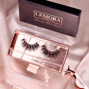 Op de afbeelding: Een close-up van een paar valse wimpers in een roze gouden doos. De wimpers zijn gelabeld "Fame" en de doos heeft de tekst "Lemora Cosmetics Luxury Mink Lashes".