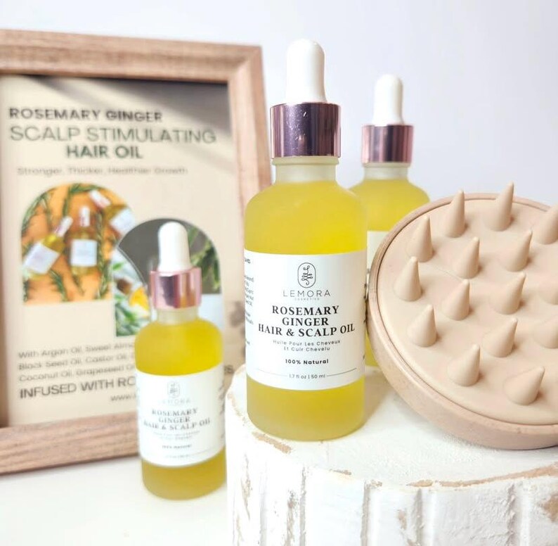 May include: Two amber glass bottles with dropper tops filled with yellow liquid. The bottles are labelled "Rosemary Ginger Hair & Scalp Oil" and are sitting on a white wooden surface. A wooden scalp massager is in the background.