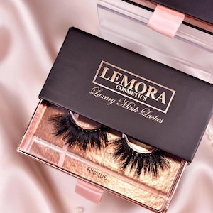 May include: A black box with gold lettering that reads "Lemora Cosmetics Luxury Mink Lashes". The box is open and reveals two pairs of false eyelashes labeled "Risqué".