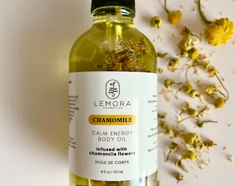 Op de afbeelding: Een doorzichtige glazen fles Lemora Cosmetics Chamomile Calm Energy Body Oil. De fles is gevuld met een gele vloeistof en bevat gedroogde kamillebloemen. Het etiket is wit met zwarte tekst die "Lemora Cosmetics Chamomile Calm Energy Body Oil Infused with chamomile flowers Huile de Corps 4 fl oz | 120 ML" luidt.