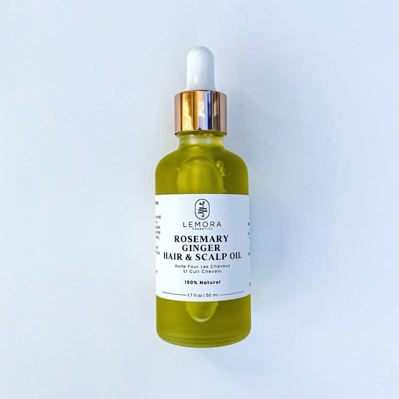 May include: A clear glass bottle with a gold cap and dropper filled with a yellow liquid. The bottle label reads "LEMORA COSMETICS ROSEMARY GINGER HAIR & SCALP OIL Huile Pour Les Cheveux Et Cuir Chevelu 100% Natural 1.7 fl oz | 50 ml"