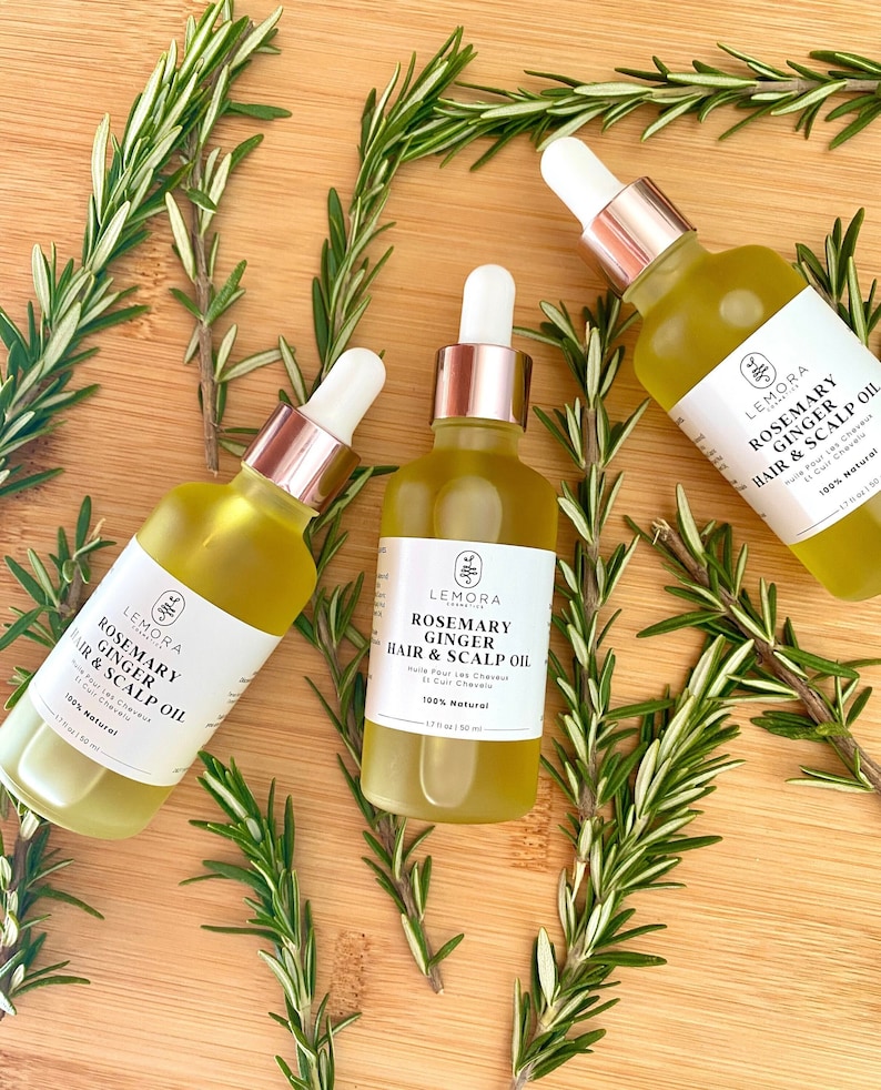 May include: Three glass bottles of rosemary and ginger hair and scalp oil with rose gold caps. The bottles are on a wooden surface with sprigs of rosemary.