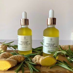May include: Two amber glass bottles with gold lids and droppers filled with a clear liquid. The bottles are labeled "Rosemary Ginger Hair & Scalp Oil" and are 100% natural. The bottles are surrounded by sprigs of rosemary and ginger root.