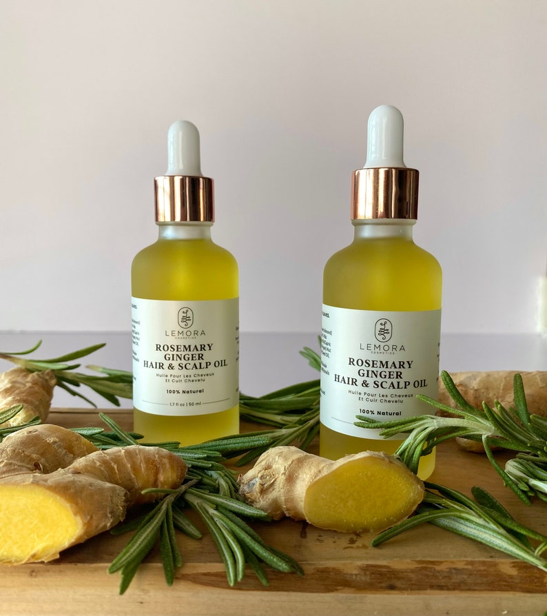 May include: Two amber glass bottles with gold lids and droppers filled with a clear liquid. The bottles are labeled "Rosemary Ginger Hair & Scalp Oil" and are 100% natural. The bottles are surrounded by sprigs of rosemary and ginger root.