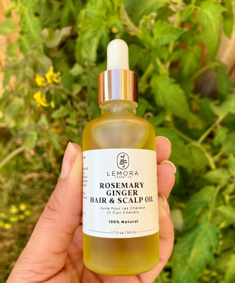 May include: A glass bottle of rosemary and ginger hair and scalp oil. The bottle is clear and has a gold cap with a dropper. The label is white with black text that reads "Rosemary Ginger Hair & Scalp Oil" and "100% Natural". The bottle is 1.7 fl oz or 50 ml.