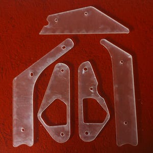 May include: A set of six clear plastic pinball flipper parts. The parts are various shapes and sizes, with rounded edges and holes for mounting.