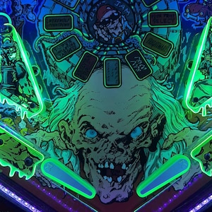 Peut inclure: Un flipper sur le thème de l'horreur, avec une grande illustration détaillée d'un visage de zombie aux yeux bleus brillants. La machine a des accents vert néon et le texte "COMPLETE CRYPT".