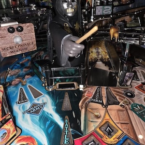 May include: A detailed view of a pinball machine featuring a figure in a black hooded robe and silver mask holding a wooden wand. The playfield has various themed elements, including text that reads "SECRET PASSAGE WHEN LIT" and "DUMBLEDORE'S ARMY".