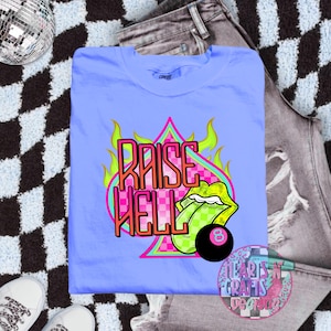 Puede incluir: Camiseta azul claro con el texto "RAISE HELL" en un diseño rosa y naranja. El diseño incluye una lengua verde, una bola negra del número 8 y llamas. La camiseta está doblada sobre una superficie a cuadros en blanco y negro.