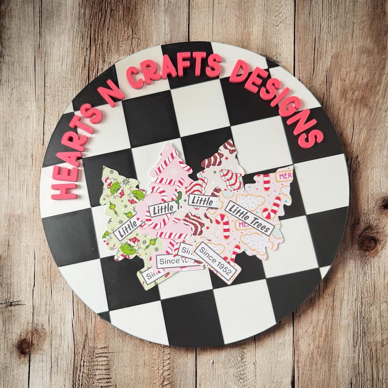 May include: A round, black and white checkered sign with the text "HEARTS N CRAFTS DESIGNS" in pink letters. The sign features Christmas tree cutouts with the text "Little Trees" and "Since 1952".