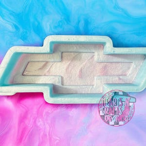 May include: A light blue Chevrolet logo mold with a pearlescent interior. The mold is set against a vibrant background of swirling blue, pink, and purple hues. The logo is a classic automotive emblem.